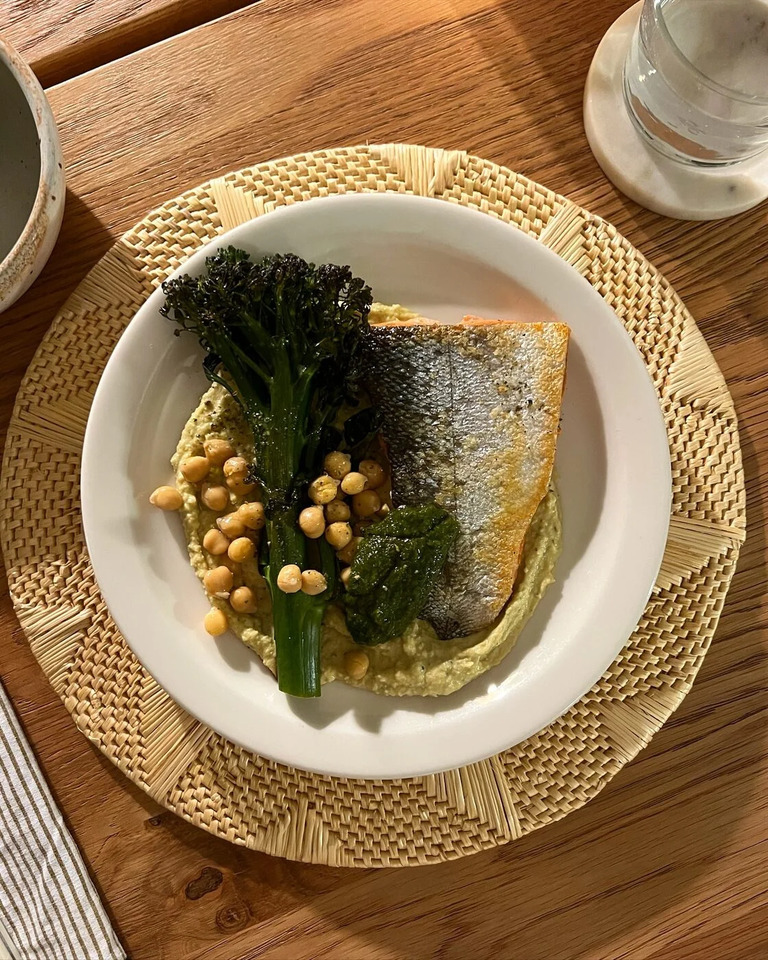 흰 접시에 구운 생선, 브로콜리니, 병아리콩, 그리고 크림 같은 소스가 담겨 있다. 접시는 라탄 재질의 매트 위에 놓여 있다.